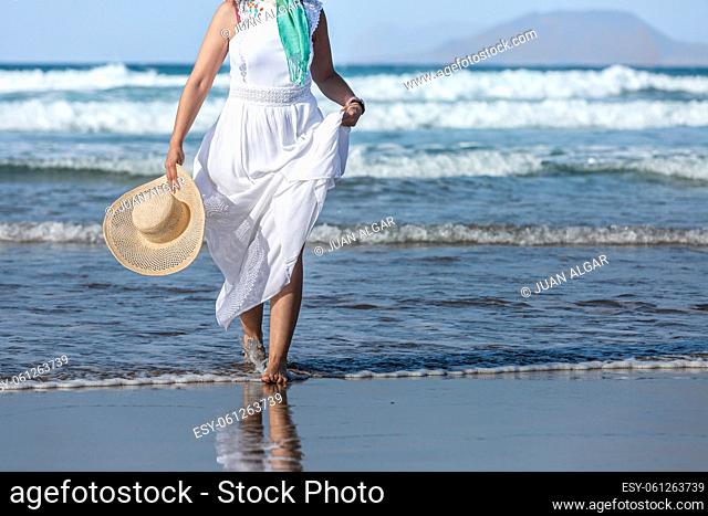 Unrecognizable barefoot female in white maxi dress carrying straw hat and walking in waves of clean sea water on summer day on resort