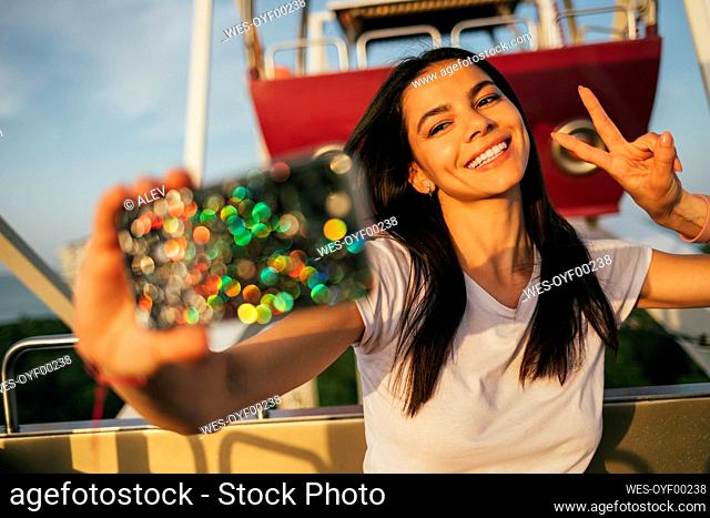 Beautiful young woman taking selfie through smart phone while gesturing peace sign on Ferris wheel