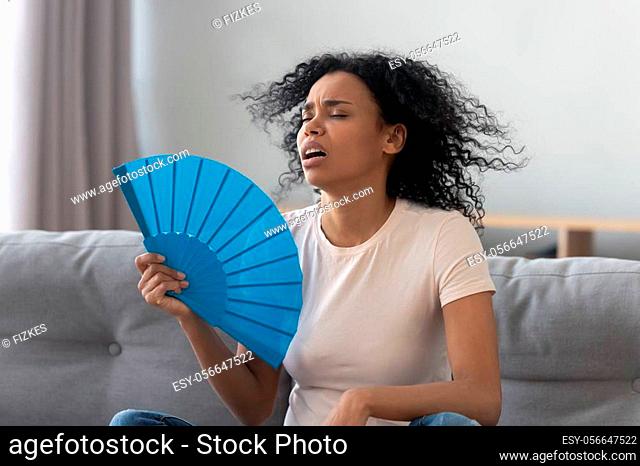 Overheated african young woman feeling hot waving fan annoyed with high temperature sit on sofa at home, stressed black girl sweating suffer from summer weather...