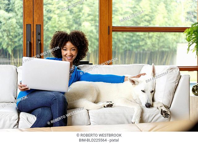 Smiling Black woman petting white dog on sofa using laptop