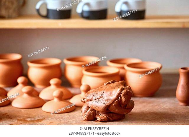 Ceramic dishware on worktop and shelves with piece of clay in front of in pottery workshop, selective focus, close-up. Plain clay mugs
