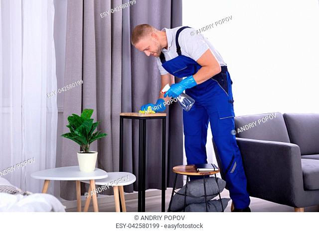 Young Male Janitor Wiping Wooden Furniture In House