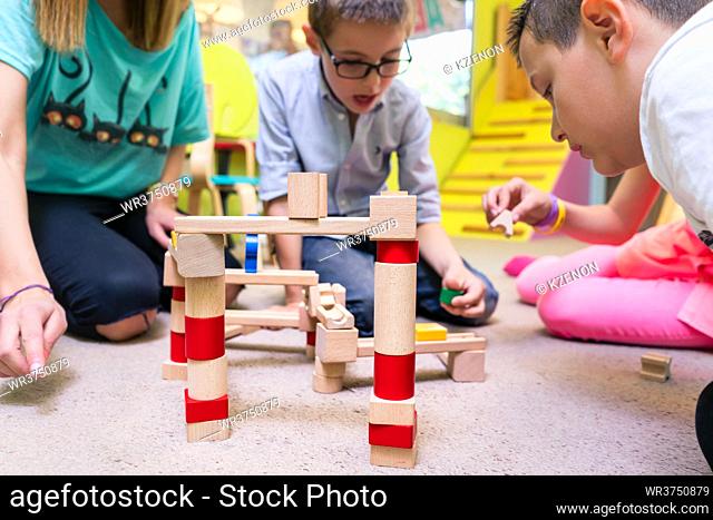 Low-angle view of a dedicated kindergarten teacher helping children with the construction of a wooden train circuit during supervised free playtime