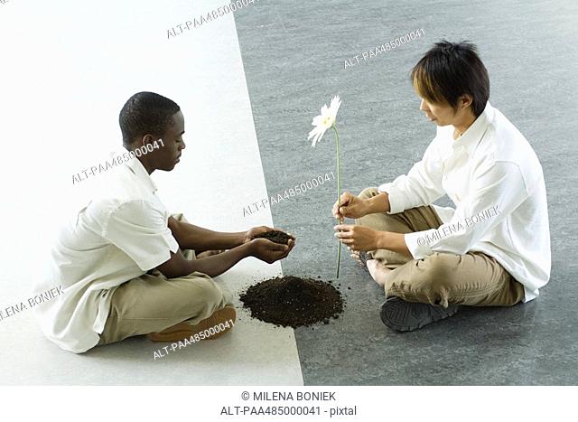 Two males sitting face to face on ground, one holding out handful of soil, the other holding out a flower