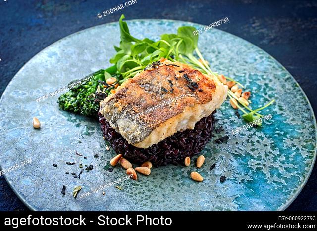Modern style traditional pan-fried skrei cod fish filet in breadcrumbs with baby broccoli, black rice and portulaca lettuce served as close-up on ceramic design...
