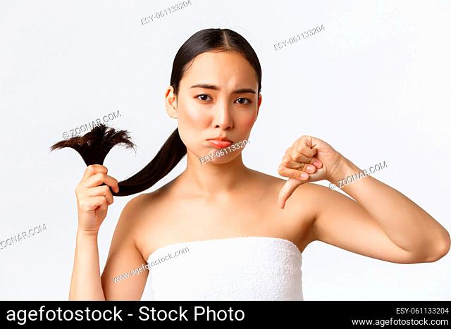 Beauty, hair loss products and hair care concept. Close-up of disappointed gloomy asian girl in bath towel, showing thumbs-up as showing split ends