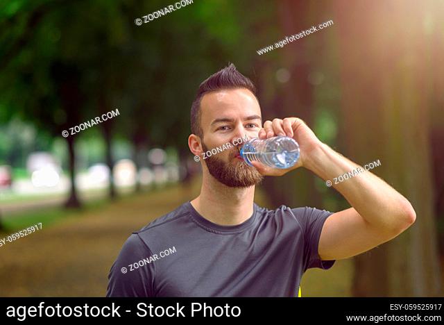 Young man with a beard drinking bottled water as he walks down a tree lined avenue in a park, close up head and shoulders