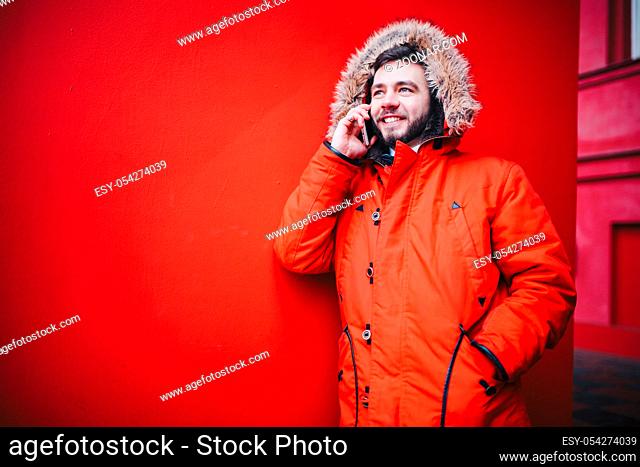 handsome young male student with a toothy smile and a beard stands on a red wall background in a bright red winter jacket with a hood with fur in winter