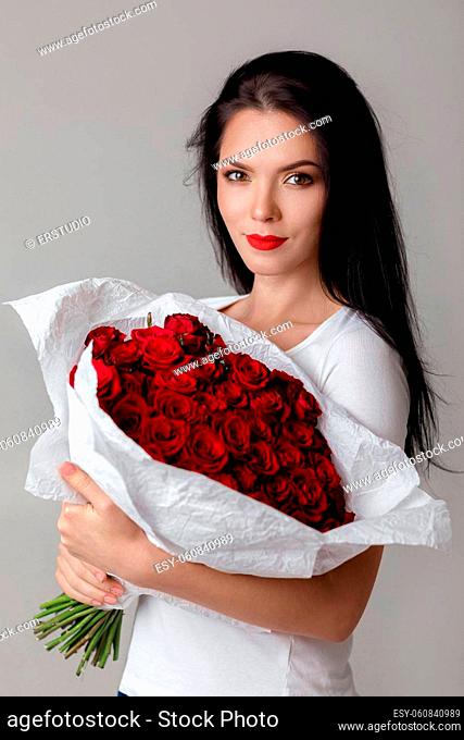 beautiful smiling young woman holding large bouquet of red roses on gray background