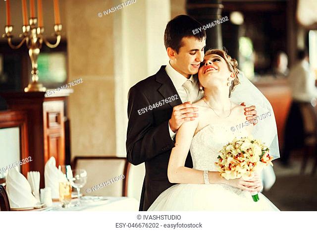 Laughing bride leans to a groom while he holds her shoulder tender