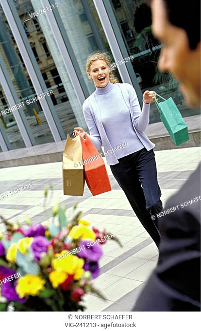 portrait, street-scene, young blond woman, wearing grey twinset and black trousers, carrying coloured paper shoppingbags is happy about a colourful bouquet of...