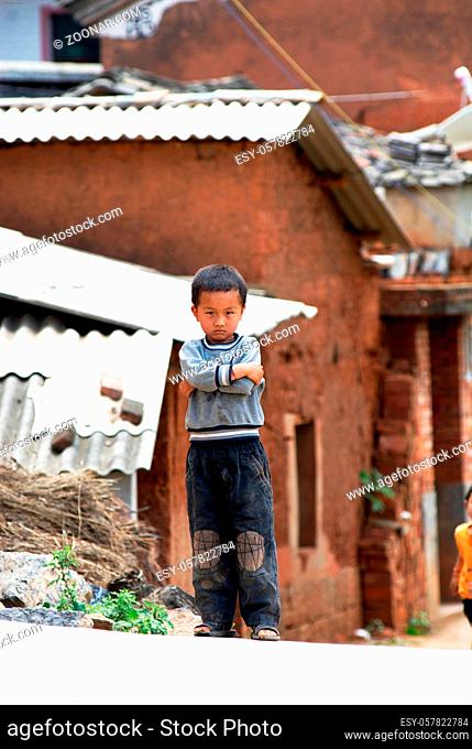 Ein chinesisches Kind mit verschraenkten Armen vor Haeusern eines chinesischen Dorfes | A chinese boy with crossed arms stands in front of houses of a chinese...