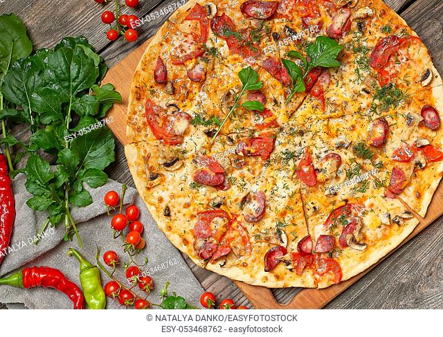 baked round pizza with smoked sausages, mushrooms, tomatoes, cheese and arugula leaves, food is cut in portions, wooden table