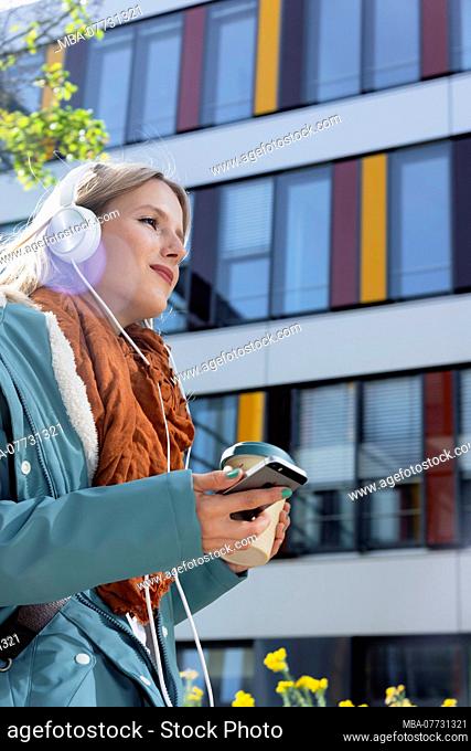Young woman with Smartphone and earphones in front of office building, half portrait