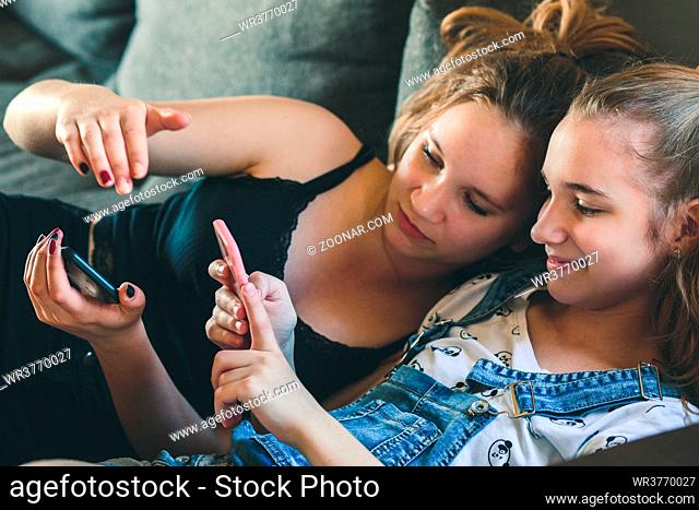 Young women using mobile phones watching music clip, texting, messaging. Teenagers using the smartphones, sitting on sofa at home