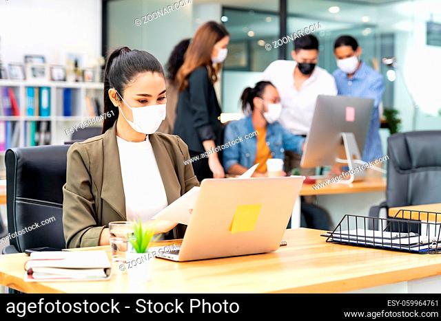 Asian office employee businesswoman wear protective face mask work in new normal office with interracial business team in background as social distance practice...