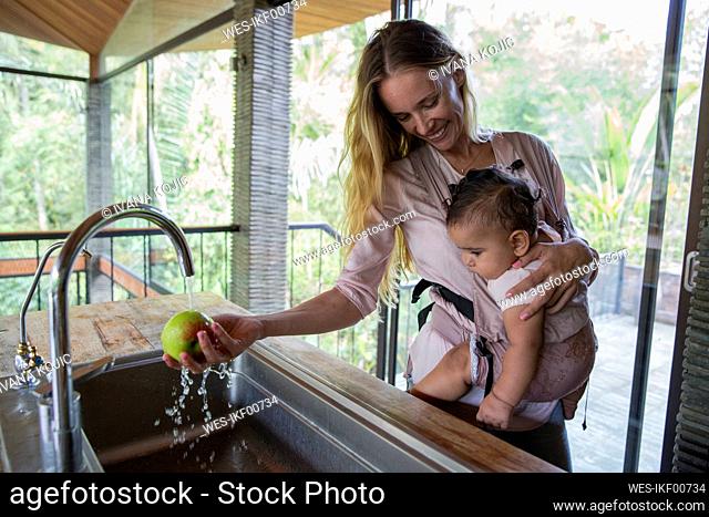 Happy woman washing apple and carrying daughter at home