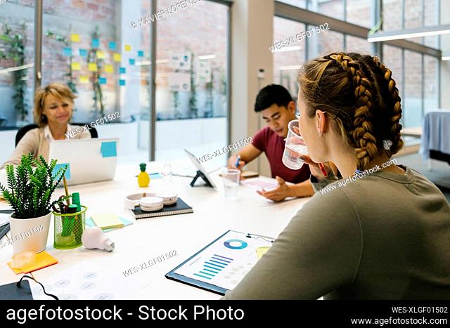 Young businesswoman drinking water at desk during meeting in coworking office