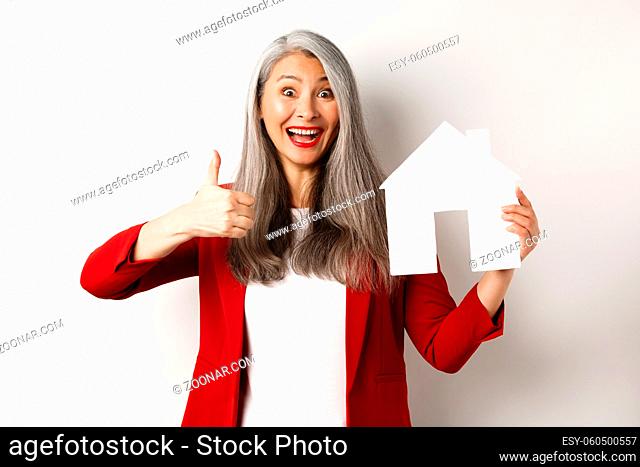 Real estate. Portrait of asian female broker showing thumb-up and paper house cutout, recommending agency to buy property, standing happy over white background
