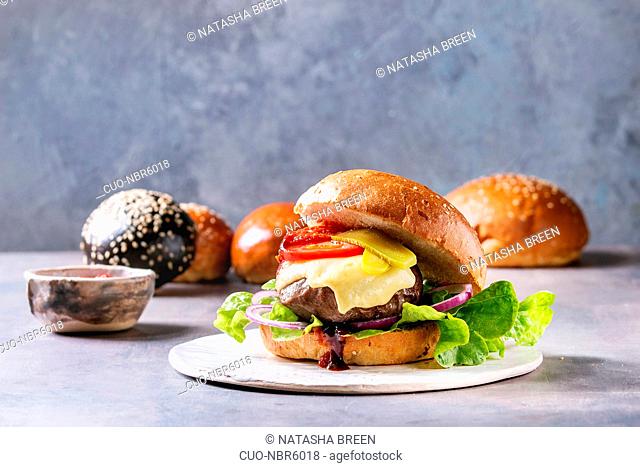 Variety of homemade classic and mini burgers in wheat and black buns with beef and veal cutlets, melted cheese and vegetables on white ceramic board over grey...