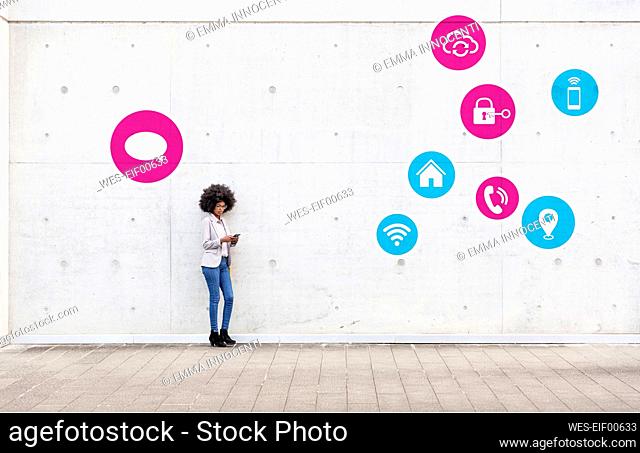Mid adult entrepreneur messaging through mobile phone while standing with icons by wall