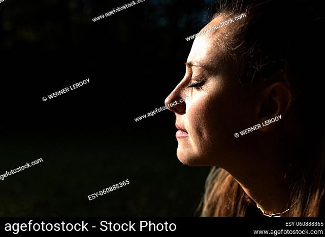 Portrait of an attractive white woman outdoors, eyes closed with the sun on her face, clair obscure