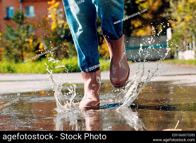 Woman wearing rain rubber boots walking running and jumping into puddle with water splash and drops in autumn rain