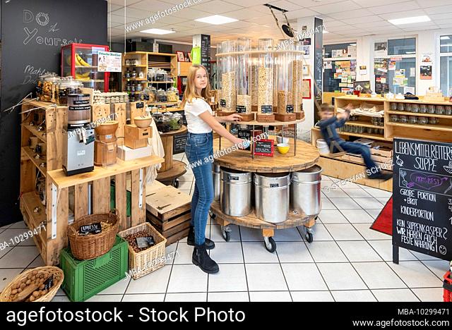 A young customer stands in front of a shelf with filling containers for muesli in an unpacked shop. A boy is swinging to the right