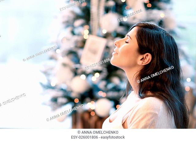 Side view portrait of beautiful brunet girl with closed eyes near beautiful decorated Christmas tree, with pleasure spending winter holidays at home