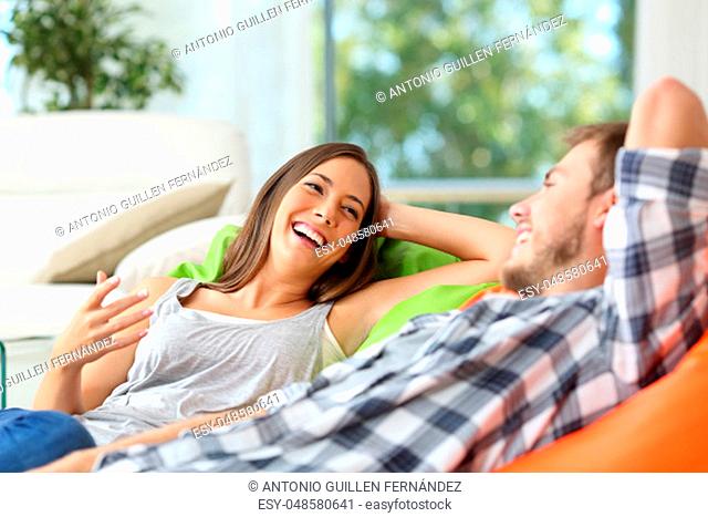 Couple or roommates talking and laughing lying on poufs in the living room at home with a window in the background