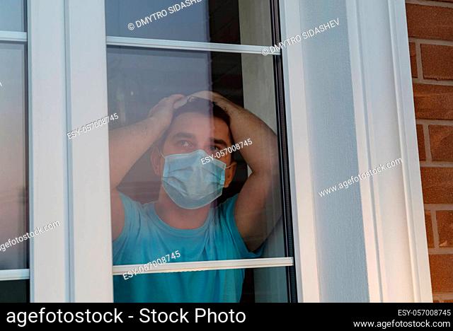 Man in a medical mask near window. Man tired of quarantine of Coronavirus. Sick sad man in medical protective face mask looking through window