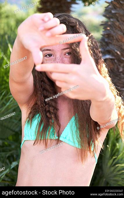 A beautiful teenage girl with freckles and wavy long hair in a bathing suit shows a frame with her hands and looks through it at the camera