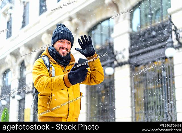 Smiling man waving hand to mobile phone while talking on video call by building during winter