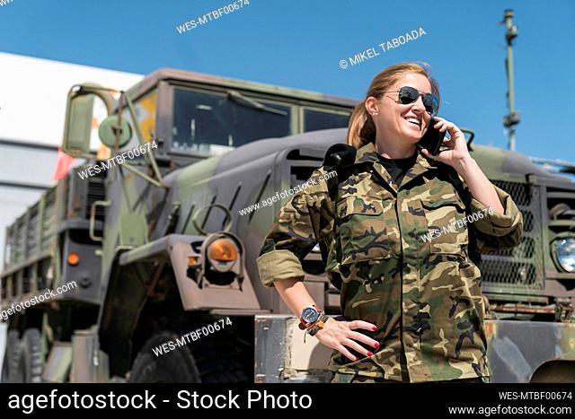 Smiling mature female army soldier talking on mobile phone while standing against truck at base on sunny day