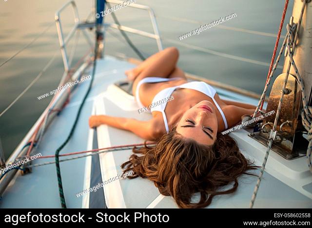 Beautiful girl with closed eyes takes sunbathe on the yacht on the background of the sea. She wears a white swimsuit. Outdoors. Horizontal