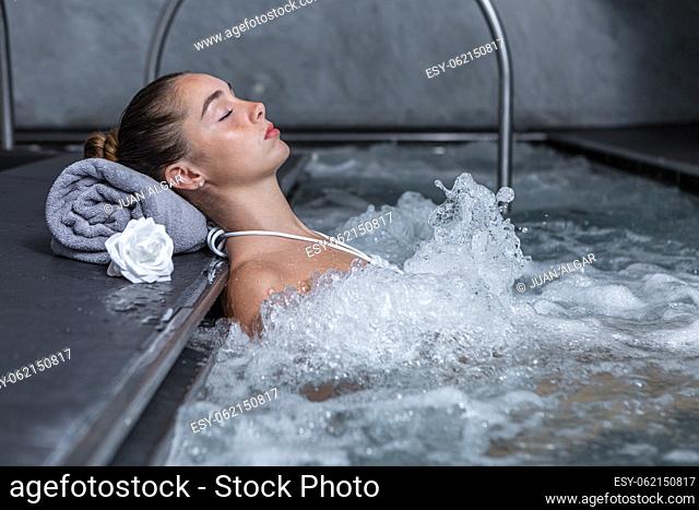 Relaxed young female with closed eyes closing eyes and holding head on rolled up towel while enjoying water massage in bubbling pool in daytime on spa resort