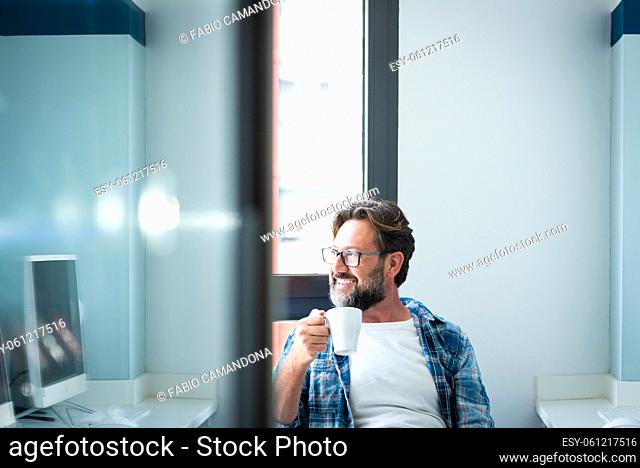 Portrait of happy man looking outside the window and smile drinking a coffee at home ot office - adult caucasian male with beard and glasses in job break...