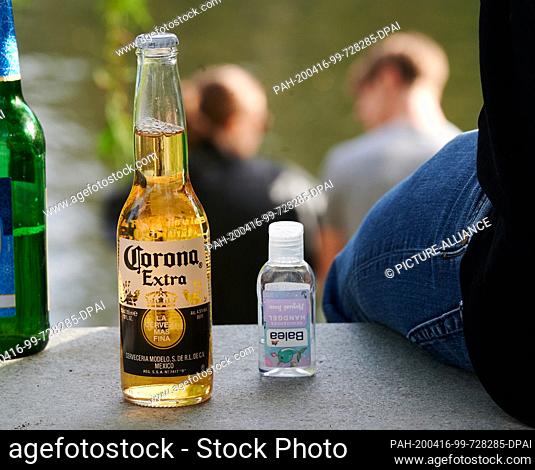 16 April 2020, Berlin: The beer Corona Extra is placed next to a bottle of cleansing hand gel next to a person. In front of them sits a couple in the sun on the...