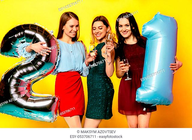 Lovely cute women in colorful dresses celebrating a birthday party, drinking a champagne, holding air balloon in form of number twenty one
