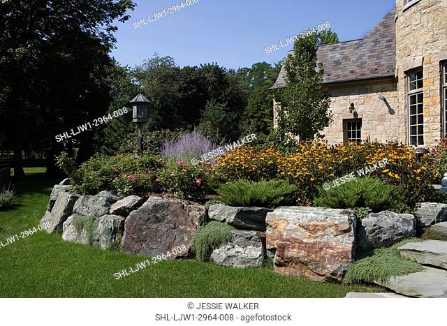 PATIOS: Backyard of French Normandy home, raised perennial border, large boulders create terrace, copper birdhouse , curve of border along grass edge, no steps
