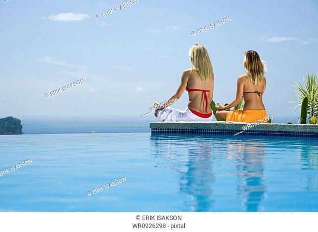 Rear view of a young woman and a mid adult woman sitting in a lotus position at the poolside