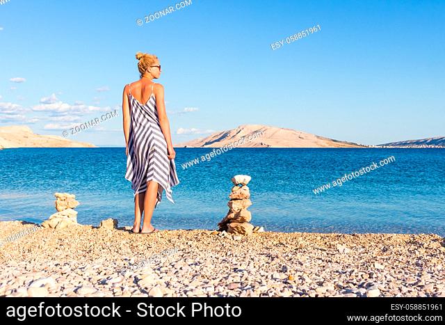 Happy carefree woman wearing beautiful striped summer dress enjoying late afternoon walk on white pabbled beach on Pag island, Croatia