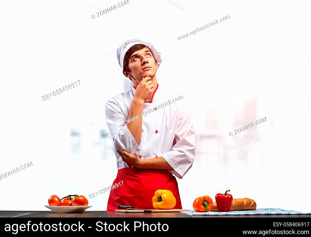 Young male chef standing with folded arms and looking up dreamily. Pensive culinary school student in white hat and red apron in light kitchen interior