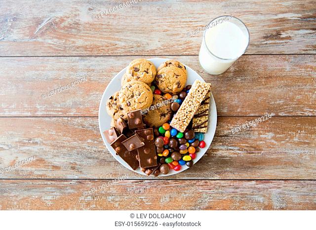 junk food, sweets and unhealthy eating concept - close up of candies, chocolate, muesli and cookies with milk glass on plate