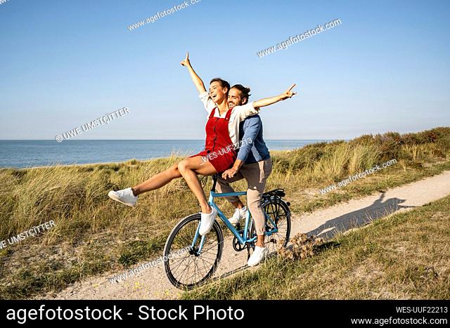 Woman with hand raised sitting on handle while enjoying bicycle ride with boyfriend against clear sky