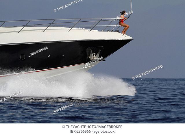 Young woman wearing a red swimsuit sitting on the railing of the bow of a moving motor yacht, French Riviera, Cote d'Azur, Mediterranean Sea, Europe