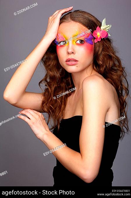 Portrait of teen girl with hand near face and orchid flower in wavy hair. Young female with unusual stylish make-up and false fashion feather eyelashes