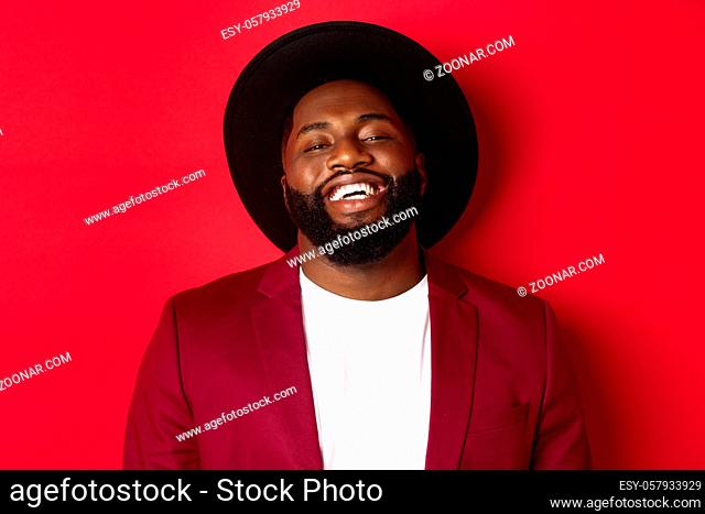 Close-up of happy handsome Black man smiling at you, looking pleased, wearing black hat and blazer, red background