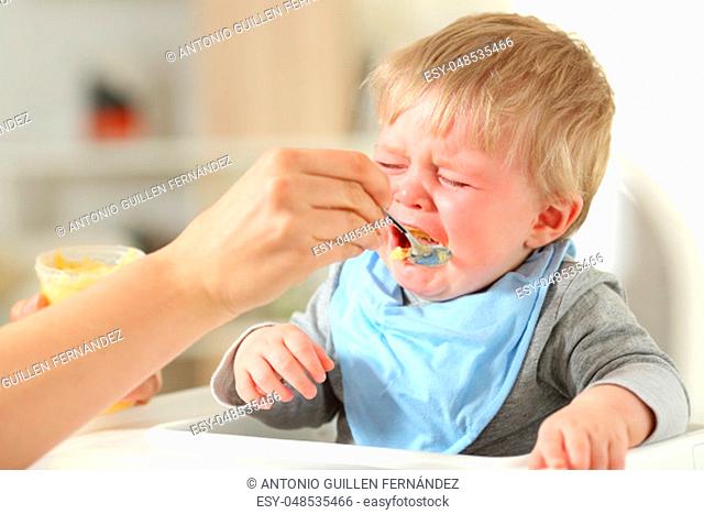 Close up of a mother hand feeding his son who is crying sitting on a high chair