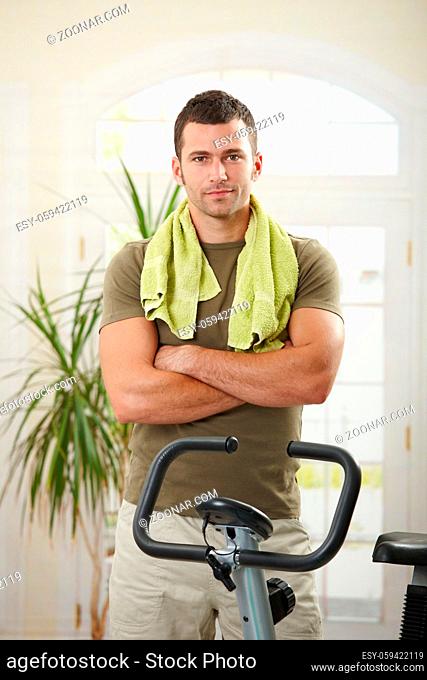 Personal trainer wearing sportswear and towel standing in living room at home with training bike, smiling